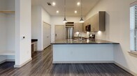 This thoughtfully designed single-floor space combines modern finishes with a functional open layout. The kitchen serves as a central highlight, featuring sleek light wood cabinetry, dark granite countertops, a textured tile backsplash, and a full suite of stainless steel appliances including a refrigerator, range, microwave, and dishwasher. A peninsula island with an undermount sink and pendant lighting overhead provides both additional workspace and a natural gathering point. Dark hardwood-style flooring flows throughout the main living areas, lending a warm yet contemporary feel. Recessed lighting illuminates the space efficiently, complementing the crisp white walls that enhance the sense of openness. The bedroom is finished with plush carpeting and features a large window dressed with horizontal blinds, allowing for natural light while maintaining privacy. The bathroom is well-appointed with large-format white tile flooring, a dark granite vanity top, light wood cabinetry, and a frameless mirror above the sink. A tiled shower enclosure adds a polished touch. The unit also includes practical elements such as a wine rack integrated into the kitchen area and ample cabinetry throughout, making efficient use of available storage. Overall, the space presents a clean, contemporary aesthetic well-suited for modern living.