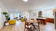 This thoughtfully designed single-floor residence showcases a seamless open-concept layout that connects the living, dining, and kitchen areas into one cohesive and airy space. Light wood flooring flows consistently throughout, contributing to a warm and inviting atmosphere. A skylight above the central living area floods the space with natural light, complementing the large windows fitted with horizontal blinds found in each room.

The kitchen is well-appointed with warm-toned wood cabinetry, white quartz countertops, stainless steel appliances including a gas range, dishwasher, and refrigerator, along with a double undermount sink positioned beneath a generous window. A peninsula with bar seating connects the kitchen to the dining area, which features a round mid-century modern dining table.

The living area offers ample space for comfortable seating and includes direct access to an outdoor area via a glass-paneled door. Adjacent rooms include a bedroom with a wall-mounted climate unit and a closet, as well as an additional flexible room suitable for a home office or secondary bedroom. The entry area is well-proportioned, featuring wall art and access to surrounding rooms. Overall, the space presents a clean, modern aesthetic with consistent mid-century design elements throughout.