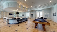 This elegantly designed single-floor amenity space blends entertainment and sophistication in a seamlessly flowing open layout. The centerpiece is a curved full-service bar with a dramatic multi-tiered glass shelf tower, a dark navy island with a white countertop, and a scalloped tile surround — all illuminated by recessed lighting set within a coffered ceiling detail above. Bar-height seating lines the counter, making it an ideal social hub. Adjacent to the bar, a regulation billiards table anchors a generous game area, complemented by views of a glazed-door arcade room nearby. The lounge area features a curated seating arrangement with a crisp white sofa, warm cognac accent chairs, bold striped area rugs, and dark wood coffee tables, all set against a striking wall of decorative paneling framing a mounted flat-screen television. Throughout the space, large framed black-and-white photographs and bold graphic artwork add a gallery-like character to the walls. Natural light streams in through multiple windows dressed with floor-length white drapery, and black-framed glass doors open to an outdoor area beyond. Underfoot, the intricate herringbone and geometric parquet hardwood flooring with dark stone inlays ties the entire space together with refined, contemporary style.