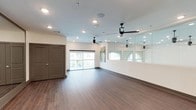 This single-floor studio space is thoughtfully designed for fitness, dance, or movement-based activities. The open floor plan provides generous room for a variety of exercises or classes, with rich hardwood-style flooring running throughout that adds warmth and durability underfoot. A defining architectural feature is the expansive floor-to-ceiling mirror wall that spans a full side of the main studio area, creating a sense of depth and allowing participants to monitor their form during activities. Multiple sleek black ceiling fans are distributed evenly across the space, ensuring consistent air circulation and comfort. Recessed lighting is positioned throughout the ceiling, offering bright, even illumination across the entire floor. Large windows along one wall flood the interior with natural light and provide pleasant views of the surrounding outdoor environment, enhancing the open and airy ambiance. A set of double doors with panel detailing provides a prominent entryway, while a secondary glass-panel door offers additional access. The clean, neutral color palette — featuring crisp white walls, light ceilings, and dark baseboards — lends the space a modern, professional aesthetic. The studio is well-suited for group fitness classes, yoga, dance rehearsals, or personal training sessions.