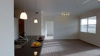 This single-story space showcases an open-concept design that seamlessly integrates the kitchen and living areas into one cohesive layout. The kitchen is anchored by a dark granite countertop that extends into a breakfast bar, illuminated by pendant lighting that hangs at varying heights, adding a layered visual appeal. A stainless steel sink and faucet are positioned along the counter, contributing to the kitchen's functional design. The transition from the kitchen into the living area is fluid, with neutral-toned carpeting covering the floor and continuing into an adjoining room visible through a doorway in the background. The living area benefits from a track lighting fixture mounted on the ceiling, providing adjustable illumination across the space. Neutral-colored walls throughout create a clean, understated backdrop, while crown molding adds a subtle architectural detail. The open doorway leading to an additional room suggests a well-connected floor plan. Natural light filters in, casting soft patterns across the carpet. Overall, the space offers a practical and cohesive environment with consistent finishes and a calm, neutral palette.