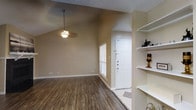 This single-story space showcases a thoughtfully designed interior that balances functionality with an open, airy feel. The main living area immediately draws the eye upward with its vaulted ceilings, which lend a sense of spaciousness and volume to the room. A ceiling fan with a light fixture hangs centrally, providing both ventilation and ambient lighting.

The flooring throughout the living area consists of durable hardwood-style planks in a warm, medium-toned finish, adding a clean and modern aesthetic. Neutral beige walls complement the flooring and contribute to a calm, cohesive color palette throughout the space.

The entryway features a solid white front door flanked by a window fitted with horizontal blinds, allowing natural light to filter in while maintaining privacy. A transition in flooring at the entry provides a distinct boundary between the main living area and the foyer.

To one side, built-in open shelving offers practical display and storage space, with tiered shelves ideal for decorative items or everyday essentials. The textured ceiling adds a subtle architectural detail consistent throughout the space. Overall, the layout feels efficient and well-organized, with a bright and inviting ambiance.