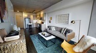 This stylish single-floor unit showcases a thoughtfully designed open-concept layout that merges the living and kitchen areas into one cohesive, functional space. The exposed concrete ceiling lends an industrial-chic character, complemented by sleek track lighting that illuminates the entire space with a warm, contemporary glow.

The living area centers around a plush gray sofa adorned with textured throw pillows, anchored by a modern marble-top coffee table set atop a rich teal area rug. The dark hardwood flooring runs throughout, providing a striking contrast against the light-toned walls and cabinetry. Curated wall art and accent lighting fixtures add personality and warmth to the space.

The kitchen is efficiently designed with clean-lined light wood cabinetry, white countertops, and stainless steel appliances, including a full-size refrigerator and built-in microwave. A compact breakfast bar with bar seating offers a casual dining option, making the kitchen as practical as it is visually appealing.

Together, the industrial architectural elements and modern furnishings create a balanced, urban ambiance that is both inviting and refined — ideal for contemporary everyday living.
