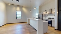 This single-floor unit showcases a contemporary design with an open-concept layout that seamlessly connects the kitchen and living areas. Soaring ceilings amplify the sense of space throughout, while light hardwood flooring runs consistently across the interior, lending warmth and cohesion to the modern aesthetic.

The kitchen is a standout feature, centered around a large white quartz island with an undermount sink and brushed brass faucet. Stainless steel appliances—including a French door refrigerator, range with stainless hood, and dishwasher—are complemented by sleek white cabinetry with dark hardware. Industrial-style pendant lighting and wall-mounted sconces add character and dimension overhead. Track lighting and a ceiling fan further illuminate the open living space, which benefits from multiple windows offering outdoor views and abundant natural light.

The bedroom is generously proportioned with soaring ceilings, black-trimmed doors, and a window with urban views. A walk-in closet with full-perimeter hanging rods provides ample storage. The bathroom features a glass-enclosed walk-in shower clad in classic white subway tile with mosaic floor tile, accented by matte black fixtures for a refined, modern finish. Bold black door frames and baseboards throughout tie the design together with a crisp, industrial-modern sensibility.