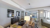 Located at 115 Amerleigh Dr in Wilmington, NC, this single-floor residence offers a thoughtfully designed open-concept layout that seamlessly connects the living and dining areas. The living room is anchored by a dark wood media console housing dual flat-screen televisions, providing a sophisticated entertainment focal point. Track lighting runs along the ceiling, offering adjustable illumination that complements the natural tones throughout the space. A tray ceiling detail adds architectural depth and visual interest to the room. Comfortable neutral-toned seating is arranged around a dark wood coffee table, set atop a vibrant geometric area rug that introduces color and texture to the hardwood floors. A pair of oversized table lamps provide warm ambient lighting, enhancing the inviting atmosphere. The space flows naturally into a dining area, where a pendant light fixture hangs above a bar-height table surrounded by sleek stools. Colorful artwork adorns the walls of the dining space, adding a curated, gallery-like quality. Decorative columns and subtle wall sconces further define the transition between spaces. The overall design blends contemporary style with warm, livable comfort throughout this well-appointed Wilmington residence.