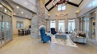 The leasing office lobby is an impressive, single-level space designed to make an immediate impact on visitors. The most striking architectural feature is the soaring vaulted ceiling, accented by dark exposed wooden beams that draw the eye upward and lend a sense of grandeur to the room. A dramatic floor-to-ceiling stacked-stone column anchors the space, serving as both a bold design statement and a functional fireplace element. Two elegant circular chandeliers with warm Edison-style bulbs hang from the peak of the vault, providing ambient lighting that complements the abundant natural light streaming in through tall, sheer-draped windows along the perimeter. The light-toned hardwood flooring runs throughout, enhancing the open, airy atmosphere. The lobby is thoughtfully divided into distinct seating areas, including a comfortable lounge zone with plush sofas, accent chairs in rich navy velvet, and a modern area rug, as well as a high-top bar seating area visible in the background — ideal for casual meetings or collaborative conversations. Neutral wall tones paired with carefully chosen furnishings in blues, grays, and gold accents create a sophisticated yet welcoming ambiance that sets a refined first impression for prospective residents.