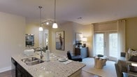 This well-designed single-floor residence offers an open-concept layout that seamlessly connects the kitchen and living areas, creating a cohesive and functional living environment. The kitchen is a standout feature, anchored by a generous granite-topped island with a double stainless steel sink, ideal for both meal preparation and casual dining. Elegant pendant lights hang above the island, complementing a track lighting system that enhances the warm, inviting ambiance of the space. Cabinetry and countertops are finished to a high standard, reflecting a modern aesthetic throughout. The open living area flows naturally from the kitchen, bathed in natural light from large windows dressed with sheer white curtains and blinds. The living space is thoughtfully arranged, featuring contemporary furnishings, including a sleek accent chair and a rustic wood side table that add character and warmth. A framed coastal artwork adorns the wall, contributing to the space's calm, curated atmosphere. Smooth, light-toned flooring runs throughout, tying the spaces together visually. The overall design balances modern finishes with comfortable, livable touches, making this residence both aesthetically refined and highly functional for everyday living.