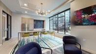 This single-floor communal lounge is thoughtfully designed with a modern aesthetic, combining warm textures and bold accents to create a vibrant yet functional gathering space. The layout is organized into several distinct zones that flow seamlessly into one another.

The lounge area features multiple seating arrangements, including plush navy blue armchairs grouped around a hammered gold accent table, complemented by a tufted sofa positioned near expansive floor-to-ceiling windows. These large black-framed windows frame dynamic urban views and flood the interior with natural light. A striking piece of contemporary wall art adds a bold focal point to one seating nook.

A linear fireplace with white herringbone tile surround is mounted beneath a large flat-screen television, anchored by a textured wood-paneled accent wall that adds warmth and depth to the space.

The kitchenette area includes light wood cabinetry, a stainless steel refrigerator, and a dark tiled backsplash serving as a dramatic backdrop. A long communal table with lime green bar-style chairs sits atop a vibrant striped area rug, tying the color palette together.

Overhead, a mid-century-inspired brass and globe chandelier provides stylish ambient lighting, reinforcing the space's polished, contemporary character throughout.