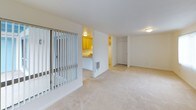This single-floor residence is part of a well-kept community distinguished by its bold blue A-frame office building, mature palm trees, manicured lawns, and colorful agapanthus plantings. A winding concrete pathway leads through the landscaped grounds to a shared pool area enclosed by a white lattice fence, creating an inviting outdoor environment.

Inside, the unit features a practical, open layout with wall-to-wall carpeting throughout the living areas and bedrooms. The kitchen is equipped with warm honey-toned wood cabinetry, a double stainless steel sink set into a laminate countertop peninsula, and a freestanding electric range with overhead exhaust. The peninsula design opens the kitchen toward the living space, promoting a connected flow between cooking and living areas.

The living room is generously proportioned, featuring vertical blind-covered sliding glass doors that allow natural light to fill the room and provide views of the exterior. A closet is conveniently located off the main living area. The bedroom is spacious and bright, with a large window dressed in vertical blinds that filter natural light across the soft carpeted floor.

The bathroom features a tub and shower combination with full tile surround in a neutral cream palette, maintaining a clean and functional aesthetic throughout. Overall, the unit offers a comfortable, move-in ready living environment within an amenity-rich community setting.