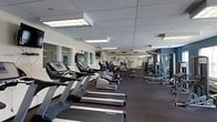 This fitness center offers a spacious, single-floor layout designed to accommodate a full range of exercise activities. The facility is outfitted with a row of True-brand treadmills along one side, providing ample cardio options for users of all fitness levels. Beyond the treadmills, the space continues into a section housing elliptical machines and additional cardio equipment, ensuring variety in aerobic training. On the opposite side of the floor, a dedicated strength training area features multi-station cable machines, weight racks, and free weight equipment, offering a comprehensive resistance training environment. The interior is finished with a drop ceiling fitted with bright fluorescent panel lighting that evenly illuminates the entire space. A wall-mounted flat-screen television is positioned for visibility from the cardio equipment, adding an entertainment element during workouts. Large windows along one wall bring in natural light and contribute to an open, airy atmosphere. The flooring consists of dark, durable carpet suited for a high-traffic fitness environment. A mirrored wall section enhances the sense of depth and allows users to monitor their form during exercise. The overall layout is clean, organized, and functional, making it a practical and well-maintained facility.