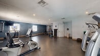 This single-floor amenity suite is thoughtfully designed to serve residents with three distinct functional areas. The spacious exercise room is the centerpiece of the facility, featuring durable rubberized flooring, recessed lighting, and a textured ceiling. It is outfitted with a variety of cardio and strength equipment, including a treadmill, stationary bike, stair climber, and a multi-station weight machine. Large windows and glass double doors flood the room with natural light, while a wall-mounted mirror enhances the sense of openness. Accent walls in a deep navy blue add a bold, motivating aesthetic to the fitness space.

Adjacent to the exercise room, the business office area provides a professional and comfortable workspace. It features warm hardwood flooring, built-in workstations with mosaic tile backsplashes, and individual computer setups separated by low partitions. A dedicated printer station and natural light from a side window make it a fully functional shared workspace. Glass double doors at the end of the office open directly to an outdoor pool area, offering a bright, welcoming connection to the exterior.

Completing the suite is a laundry room, conveniently accessible for residents. Together, these spaces create a comprehensive, well-appointed amenity facility.