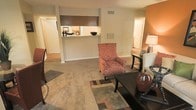 This single-story space features an open-concept layout that connects the living area to the kitchen through a convenient pass-through counter, enhancing the sense of flow between rooms. The living area is finished with wall-to-wall carpeting and anchored by a patterned area rug, creating a comfortable and inviting atmosphere. Warm neutral wall tones are accented by a bold terracotta-orange feature wall, adding visual depth and character to the space. The pass-through kitchen counter provides a functional serving and prep surface while maintaining visual connectivity to the living area. Overhead, a ceiling vent ensures consistent climate control throughout. A hallway visible in the background suggests additional rooms beyond the main living space, offering practical separation of living and private areas. The layout is well-suited for everyday living, with ample room for seating arrangements centered around a dark wood coffee table. Overall, the space combines functional design with warm, earthy tones to create a cozy and cohesive interior environment.