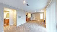 This single-floor residence offers a practical and well-organized layout designed for comfortable everyday living. The main living area is spacious and open, finished with neutral-toned carpet that extends throughout, contributing to a warm and cohesive atmosphere. Recessed lighting in the ceiling provides consistent, even illumination across the space, while a window allows natural light to brighten the interior.

The kitchen is positioned in an open-concept arrangement, visible from the main living area and anchored by a peninsula-style breakfast bar with warm wood-toned cabinetry. This layout encourages a connected flow between cooking and living spaces. Appliances and upper cabinetry are neatly integrated, making the kitchen both functional and visually tidy.

The living area also features a thermostat panel and a ventilation access panel mounted on the wall, indicating thoughtful climate control infrastructure. A set of double doors along the interior wall suggests generous storage or closet space, adding to the home's practical appeal. Smooth, light-gray walls complement the soft carpeting and white trim throughout, lending the space a clean, understated aesthetic. A side door entry point adds additional accessibility to the layout. Overall, the space presents a well-maintained, move-in-ready interior with a straightforward and functional design.