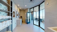 This upscale pet spa and grooming facility occupies a single floor of a high-rise building, combining sleek modern design with practical, pet-focused amenities. The entrance area welcomes visitors with polished light-toned flooring, recessed ceiling lighting, and a series of large-format framed photographs of dogs displayed along the walls, setting a warm yet sophisticated tone throughout.

The grooming area is thoughtfully equipped with a deep built-in bathing tub, neatly stocked shelving units filled with fresh towels, wall-mounted grooming tools, and a paw-print padded bench for pet handling. Cabinetry in a wood-grain finish provides ample storage, while the clean, light-colored surfaces ensure a hygienic and organized environment. A utility sink with upper and lower cabinetry is conveniently positioned near the entry, offering additional workspace for grooming professionals.

The lounge area is a standout feature, enclosed by expansive floor-to-ceiling glass walls that frame sweeping panoramic views of the surrounding cityscape and waterway beyond. Sleek black lounge seating and sculptural dog statues add an artistic touch, reinforcing the space's dedication to canine culture. Dark-framed glass doors connect the various zones, maintaining visual openness while defining each functional area. Overall, the space blends high-end finishes with purposeful design, creating a refined environment for pets and their owners alike.