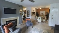 This single-floor residence offers a thoughtfully designed open-concept layout that connects the living, dining, and kitchen areas into one cohesive space. The living area is anchored by a tile-surround fireplace and features warm, neutral carpeting, a ceiling fan, and a wall-mounted flat-screen television. Stylish patterned accent chairs and a glass coffee table contribute to the contemporary aesthetic, while a decorative oval mirror adds visual depth to the charcoal accent wall.

Just beyond the living area, a compact dining space accommodates a round glass-top table surrounded by cream upholstered chairs, creating an intimate setting for meals. Track lighting overhead highlights this area effectively. A pass-through or bar-height counter connects the dining space to the kitchen, which is visible in the background and features dark cabinetry for a bold contrast against the lighter walls.

The kitchen is equipped with dark-finished cabinetry and appears well-organized, offering functional workspace. Throughout the residence, the interplay of warm earth tones, accent walls in gray and tan, and carefully chosen furnishings create a modern yet inviting ambiance.