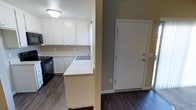 This single-story space showcases a functional and clean interior design. The kitchen is a standout feature, equipped with white cabinetry that extends to the upper and lower walls, providing ample storage. A peninsula with a white countertop extends into the space, incorporating a stainless steel sink and creating a natural division between the kitchen and the adjacent entry area. A black range is visible along the kitchen wall, complementing the overall color scheme. The flooring throughout is a dark hardwood-style material, offering a rich contrast to the white cabinetry and light-colored walls. The walls are finished in a neutral taupe tone, contributing to a calm and cohesive ambiance. A white interior door near the kitchen area leads to an adjoining room or entry point, framed with clean white trim that matches the cabinetry. Ceiling-mounted lighting provides overhead illumination, and a window above the sink allows for natural light to filter into the kitchen. The overall layout is straightforward and efficient, making practical use of the available space.
