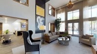 This stylish lounge space makes an immediate impression with its dramatic double-height ceilings, creating an expansive, airy atmosphere that draws the eye upward. The architectural design incorporates a striking dark wood accent wall, which serves as a backdrop for a curated gallery of bold, contemporary artwork in yellow, white, and navy tones, adding visual depth and artistic character to the space.

The seating area is thoughtfully arranged for both comfort and aesthetics, featuring a plush taupe sofa paired with a coordinating ottoman and an oversized dark charcoal wingback chair. A sculptural stone coffee table anchors the seating arrangement, while a tall, ornate floor lamp with an intricate base adds ambient lighting and design interest.

Sheer linen drapery lines the floor-to-ceiling windows and glass doors, which flood the interior with natural light and offer direct views of a pool area and lush palm trees beyond. A woven chandelier overhead adds warmth and texture to the high ceiling above.

Additional spaces are visible through open doorways, suggesting a larger amenity facility. Tall indoor bamboo plants bring a natural, organic element to the polished, contemporary interior.