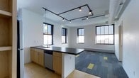 This contemporary single-floor space showcases a thoughtfully designed open-plan layout that seamlessly blends functional and social areas. The kitchen stands out as a central feature, equipped with a dark countertop island, light wood cabinetry, a stainless steel sink, and a built-in dishwasher — offering ample workspace and storage. A pendant light fixture adds a touch of warmth above the kitchen area.

Overhead, a striking black track lighting system runs across the white ceiling, providing versatile, adjustable illumination throughout the space. The clean white walls and crisp trim work contribute to a bright, modern aesthetic.

Multiple large black-framed windows line the walls, inviting an abundance of natural light and offering views of the surrounding urban landscape. The flooring transitions from smooth hardwood in the kitchen zone to a patterned carpet in shades of blue and yellow in the open living or lounge area, adding visual interest and defining the separate functional zones.

A baseboard heating unit is visible along the far wall, ensuring year-round comfort. The overall ambiance is clean, airy, and versatile — well-suited for a variety of uses, from a working studio to a collaborative or residential space.