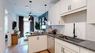 This thoughtfully designed single-floor residence showcases a clean, contemporary aesthetic with an open-concept layout that connects the kitchen and living area seamlessly. Light wood-look flooring runs throughout, lending warmth and continuity to the space.

The kitchen is a standout feature, appointed with crisp white shaker cabinetry, dark granite countertops, and a striking herringbone tile backsplash. A full suite of stainless steel appliances — including a range, built-in microwave, dishwasher, and refrigerator — is complemented by a matte black faucet and hardware, adding a modern contrast. Pendant lights with black shades hang above the peninsula, which opens directly into the living area.

The living room is bright and inviting, anchored by a large window with floor-to-ceiling drapes that allow natural light to pour in. A ceiling fan overhead and a teal accent wall provide a pop of color and character, while the open sightlines create a sense of spaciousness.

The bathroom is clean and functional, featuring large-format tile in the tub and shower surround, built-in open shelving for storage, and consistent light flooring. The overall design of the unit balances modern finishes with a warm, livable atmosphere throughout.