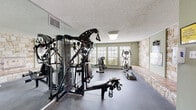 Located at 7771 Joe Newton Rd in San Antonio, Texas, this dedicated fitness center offers a well-equipped, single-floor workout facility designed to support a comprehensive exercise routine. The space is defined by a striking natural limestone accent wall that runs along one side of the room, complemented by a built-in tiled bench — adding a distinctive Texas character to the functional environment. Soft sage-green walls and a textured ceiling contribute to a clean, focused atmosphere throughout.

The fitness floor features a multi-station cable strength machine as its centerpiece, supported by a weight bench and an assortment of additional strength-training equipment. Cardio options include an elliptical trainer and a stationary bike, positioned near a bank of large windows fitted with horizontal blinds that allow ample natural light to filter into the space while maintaining privacy. Floor-to-ceiling mirrors line one wall, visually expanding the room and allowing users to monitor their form during workouts.

Durable rubber flooring covers the main exercise area, providing both safety and comfort underfoot. The room is well-lit with overhead flush-mounted lighting and includes proper ventilation. Safety equipment such as a fire extinguisher is also present. The space is accessed through a dedicated entry door, reinforcing its role as a self-contained, purpose-built fitness facility.