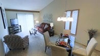 This single-floor space offers a seamlessly connected living and dining area designed for both comfort and functionality. The open-plan layout is anchored by soaring vaulted ceilings that lend an airy, expansive feel to the interior. Large windows dressed with vertical blinds line the walls, flooding the space with abundant natural light and contributing to the overall sense of openness.

The dining area is defined by an elegant glass-top table surrounded by upholstered cream chairs, set beneath a contemporary multi-bulb chandelier that serves as a refined focal point. Decorative table settings add a touch of warmth and character to the space.

Adjacent to the dining area, the living room features plush, neutral-toned carpeting underfoot and is furnished with a rich brown sofa accented by patterned throw pillows. A round glass coffee table and coordinating accent chairs round out the seating arrangement, creating an inviting area for relaxation. A tasteful wall accent piece in an organic, branch-like motif adds a subtle artistic element to the shared space.

The cohesive color palette of warm neutrals and earthy tones throughout ties the two areas together harmoniously, resulting in a welcoming and well-appointed interior.