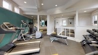 This fitness center offers a thoughtfully designed workout environment spread across a single level. The space is defined by crisp white walls accented by bold teal panels, creating an energetic yet clean atmosphere. Large windows positioned along the upper portions of the walls invite abundant natural light throughout the facility, enhancing the open feel of the interior.

The main workout area is well-appointed with a variety of cardio equipment, including multiple treadmills, an elliptical trainer, a stationary bike, and a rowing machine. A wall-mounted flat-screen television is positioned for easy viewing during cardio sessions. A dedicated free weights section features a full dumbbell rack alongside an adjustable workout bench, while a multi-functional cable machine provides options for comprehensive strength training.

Full-length mirrors line one wall, amplifying the sense of space and allowing users to monitor their form during exercise. The flooring consists of large-format gray tiles accented with teal and yellow geometric inlays, adding visual interest underfoot. Recessed lighting provides consistent illumination throughout. A white railing delineates the entry area, which also includes a wall-mounted sanitizing station and posted facility guidelines, contributing to a clean and organized environment.