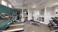 This fitness center offers a thoughtfully designed workout environment spread across a single level. The space is defined by crisp white walls accented by bold teal panels, creating an energetic yet clean atmosphere. Large windows positioned along the upper portions of the walls invite abundant natural light throughout the facility, enhancing the open feel of the interior.

The main workout area is well-appointed with a variety of cardio equipment, including multiple treadmills, an elliptical trainer, a stationary bike, and a rowing machine. A wall-mounted flat-screen television is positioned for easy viewing during cardio sessions. A dedicated free weights section features a full dumbbell rack alongside an adjustable workout bench, while a multi-functional cable machine provides options for comprehensive strength training.

Full-length mirrors line one wall, amplifying the sense of space and allowing users to monitor their form during exercise. The flooring consists of large-format gray tiles accented with teal and yellow geometric inlays, adding visual interest underfoot. Recessed lighting provides consistent illumination throughout. A white railing delineates the entry area, which also includes a wall-mounted sanitizing station and posted facility guidelines, contributing to a clean and organized environment.