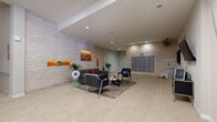 Located on Ventura Boulevard in the Encino neighborhood of Los Angeles, this building lobby and hallway space offers a polished, contemporary environment designed to make a strong first impression. The main lobby is generously proportioned, finished with large-format light stone tile flooring that extends throughout and contributes to a clean, cohesive aesthetic.

The focal point of the lobby is a striking textured stone accent wall featuring warm, backlit display niches in deep orange — a standout architectural detail that adds visual warmth and a gallery-like quality to the space. Recessed lighting illuminates the area evenly, complementing the modern seating arrangement of sleek charcoal upholstered chairs, a glass-top coffee table, and a plush area rug.

A dedicated mailbox area lines one wall, clad in matching stacked stone tile and housing a full bank of individual resident mail compartments. Adjacent to the lounge, a secondary hallway leads to the building entrance, featuring floor-to-ceiling glass doors that invite natural light deep into the space and provide clear sightlines to the exterior.

The connecting hallways are bright and well-maintained, with recessed ceiling lights, neutral wall tones, and framed artwork adding character. A resident workspace or management area with a mounted monitor and furnishings rounds out the functional offerings of this well-designed lobby.
