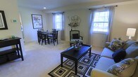 This single-floor space offers a comfortable and well-appointed layout that seamlessly blends living and dining areas into one cohesive environment. The living area is anchored by a dark wood coffee table set atop a bold geometric-patterned area rug, complemented by a light-toned sofa and a decorative accent chair upholstered in a green patterned fabric. Neutral carpeting runs throughout, adding warmth and a sense of continuity across the space. The dining area features a dark wood table paired with matching chairs, positioned near a framed wall art piece depicting a bicycle with flowers, lending a charming, curated feel to the corner. Multiple double-hung windows dressed with sheer white curtains and coordinating drapery panels flood the space with abundant natural light, creating a bright and airy atmosphere. The walls are finished in a soft, warm neutral tone that enhances the sense of openness. Decorative touches, including a sculptural metal wall piece and a floral arrangement, add character and visual interest throughout. Recessed lighting provides additional illumination, ensuring the space is well-lit at all hours. Overall, this space presents a clean, inviting aesthetic suited for both relaxed everyday living and casual entertaining.