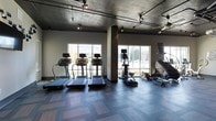 This single-floor fitness center offers a well-appointed workout environment designed with both functionality and aesthetics in mind. The space features an exposed concrete ceiling with industrial-style track lighting, lending a contemporary, urban feel to the interior. Multiple large windows line the walls, allowing abundant natural light to brighten the gym floor and providing views of the surrounding outdoor scenery.

The cardio area is well-equipped, housing a row of treadmills along the windowed wall, complemented by elliptical trainers and stair climbers positioned nearby. Each machine is outfitted with digital display screens, supporting a variety of workout programs. Structural columns add to the industrial character of the space while subtly defining equipment zones.

The flooring consists of patterned, multi-toned gym tiles that provide durability and traction underfoot. Track lighting mounted on exposed ceiling rails ensures the entire room is evenly illuminated. The neutral color palette of grey, white, and muted tones throughout the space contributes to a clean, focused atmosphere well-suited for exercise.