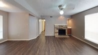 This single-story home offers a practical and comfortable layout with a mix of hardwood-style and carpeted flooring throughout. The living area serves as the heart of the home, showcasing a classic brick fireplace as its focal point, complemented by dark laminate flooring, a ceiling fan with light fixture, and a textured ceiling. A hallway connects the living space to the rest of the home, while a pass-through area adjacent to the fireplace provides a glimpse into the kitchen. The kitchen is efficiently designed with warm wood cabinetry, white appliances including an electric range and full-size refrigerator, and light countertops. The bedrooms are carpeted for comfort, each featuring ceiling fans, neutral-toned walls, and large windows fitted with blinds that allow for natural light. The primary bathroom includes a vanity with wood cabinetry, a tub-shower combination, and direct access to a spacious walk-in closet outfitted with dual hanging rods and a central shelving unit. A second bathroom also features a tub-shower combo. The entry foyer, visible from the main living area, is bright and welcoming with a windowed front door. Throughout, the home maintains a consistent, neutral color palette that creates a clean and cohesive atmosphere.