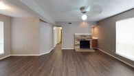 This single-story home offers a practical and comfortable layout with a mix of hardwood-style and carpeted flooring throughout. The living area serves as the heart of the home, showcasing a classic brick fireplace as its focal point, complemented by dark laminate flooring, a ceiling fan with light fixture, and a textured ceiling. A hallway connects the living space to the rest of the home, while a pass-through area adjacent to the fireplace provides a glimpse into the kitchen. The kitchen is efficiently designed with warm wood cabinetry, white appliances including an electric range and full-size refrigerator, and light countertops. The bedrooms are carpeted for comfort, each featuring ceiling fans, neutral-toned walls, and large windows fitted with blinds that allow for natural light. The primary bathroom includes a vanity with wood cabinetry, a tub-shower combination, and direct access to a spacious walk-in closet outfitted with dual hanging rods and a central shelving unit. A second bathroom also features a tub-shower combo. The entry foyer, visible from the main living area, is bright and welcoming with a windowed front door. Throughout, the home maintains a consistent, neutral color palette that creates a clean and cohesive atmosphere.