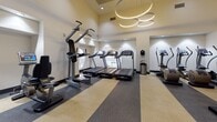 Located at 8000 West Dr in North Bay Village, Florida, this contemporary fitness center is thoughtfully designed to support a full range of workout activities across a single, well-organized floor. The facility makes a strong visual impression with its bold teal accent walls, striped multi-tone carpeted flooring, and vibrant framed artwork throughout, creating an energetic yet polished atmosphere.

The main fitness floor is divided into distinct zones, beginning with a spacious cardio area equipped with treadmills, ellipticals, and cycling machines beneath a striking circular chandelier. Nearby, a dedicated strength training zone offers a cable machine, free weights, barbells, and benches, with teal accent walls and mirrored surfaces enhancing the sense of space.

A glass-enclosed group exercise studio provides a dedicated setting for spin classes or guided workouts, fitted with stationary bikes and a large display screen, with natural light filtering in through windows overlooking lush greenery. An open stretching and functional training area features suspension trainers and stability equipment along the perimeter.

The facility also includes a relaxed lounge area with comfortable woven chairs arranged around a small table, offering a welcoming space to rest or consult with a trainer. Motivational typography accents the walls, reinforcing an atmosphere centered on wellness, focus, and performance.