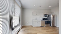 This single-floor unit presents a clean, contemporary design throughout, with light hardwood-style flooring and neutral gray-toned walls creating a cohesive, modern aesthetic across all rooms. The open layout connects the living area to the kitchen, maximizing the sense of space and encouraging functional flow.

The kitchen is well-appointed with white shaker-style cabinetry, stone-look countertops, and a stainless steel undermount sink with a pull-down faucet. Stainless steel appliances include a gas range, built-in microwave, and dishwasher, providing a fully equipped cooking space with a polished, uniform finish.

The living area is a standout feature, anchored by a large floor-to-ceiling sliding glass window wall fitted with horizontal blinds. This expansive glazing draws in abundant natural light while offering views of the surrounding streetscape, giving the room an open, airy character.

A dedicated bedroom offers a quiet, simply finished space with the same light hardwood flooring and neutral walls. The bathroom includes a full tub-shower combination with a white surround, chrome fixtures, and coordinating flooring, completing the unit's consistent modern palette. Recessed lighting throughout adds a refined, unobtrusive illumination to the overall space.