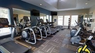 This fitness center offers a well-appointed workout environment designed to support a full range of exercise routines. The space is outfitted with multiple treadmills and elliptical cross-trainers, all equipped with individual digital displays, providing a robust cardio zone for users of varying fitness levels. A dedicated free weights area features a neatly organized dumbbell rack positioned near large windows, allowing ample natural light to fill the room and offering views to the outdoors.

The interior design strikes a balance between functionality and modern aesthetics. Deep teal accent walls contrast with neutral tones on the ceiling and trim, creating an energizing yet comfortable atmosphere. Large mirrors are mounted along one wall, enhancing the sense of space and allowing for proper form checks during workouts. Patterned carpet tiles cover the floor, adding texture and visual interest while providing a practical surface underfoot.

Overhead, recessed lighting complements the natural light from the multiple windows lining the exterior wall. Wall-mounted flat-screen televisions are positioned for easy viewing from the cardio equipment, supporting an engaging workout experience. The room's raised ceiling tray detail adds an architectural touch, contributing to the overall polished and welcoming feel of this fitness facility.