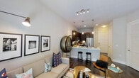 This single-floor residence features an open-concept layout that seamlessly connects the living area and kitchen, creating a cohesive and functional living environment. The living space is anchored by a neutral-toned sofa accented with vibrant throw pillows, complemented by a patterned area rug and curated wall art, lending the room a contemporary yet welcoming character. Dark hardwood floors run throughout, providing a rich contrast to the bright white walls and ceiling.

The kitchen is designed with dark cabinetry, stainless steel appliances, and a white countertop island that doubles as a casual dining area, complete with sleek bar stools. Pendant lights hang above the island, adding warmth and visual interest, while a track lighting system overhead ensures the entire space is well-illuminated. Recessed lighting further enhances the bright, airy atmosphere throughout.

The ceilings are notably tall, amplifying the sense of openness and space. The overall design aesthetic is modern and urban, with thoughtful attention to detail in the fixtures and finishes, creating an inviting and sophisticated environment suitable for both relaxing and entertaining.