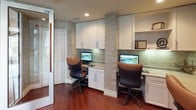 This business center offers a thoughtfully designed workspace that balances functionality with refined aesthetics. The entrance is defined by striking glass-paneled doors featuring geometric, angular patterns that lend a contemporary architectural character to the space. Rich hardwood flooring flows throughout, contrasting elegantly with the stone tile found near the entryway.

The main work area is equipped with multiple built-in workstations, each featuring a dedicated computer terminal, ergonomic seating, under-cabinet lighting, and generous white cabinetry for storage above and below. Textured stone accent walls add warmth and visual interest between the workstation bays, contributing to an atmosphere that feels both professional and inviting.

Natural light filters into the space through large windows adorned with the same distinctive geometric glass detailing seen at the entrance, offering views to an adjacent corridor and conference area. Recessed ceiling lighting provides consistent, well-distributed illumination throughout.

The overall layout is efficient and clearly organized, with signage indicating business center rules and policies. The combination of warm wood tones, neutral stone textures, and modern furnishings creates an environment well-suited for focused work, making this business center a practical and visually cohesive amenity space.
