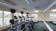 This single-floor fitness center offers a dedicated space for cardiovascular exercise and general wellness. The room is lined with a generous row of cardio equipment, including elliptical trainers, treadmills, and additional exercise machines, all arranged to maximize usable floor space. Large windows span the walls, flooding the interior with natural light while providing views of the surrounding outdoor greenery, creating an open and energizing atmosphere during workouts. The light-toned walls and green carpeted flooring give the space a clean, practical aesthetic. Multiple flat-screen televisions are mounted throughout the room, allowing users to stay entertained or informed during their sessions. Overhead, tube-style lighting fixtures supplement the natural light, ensuring the space remains well-lit at all times. A wall-mounted air conditioning unit contributes to a comfortable climate year-round. The layout is thoughtfully organized, with equipment arranged in parallel rows to facilitate easy movement and access. Overall, this fitness center is a functional and inviting space, well-suited to support a range of cardio-focused exercise routines.