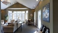 This single-floor space is defined by its striking vaulted ceilings, which rise dramatically to create an airy, open atmosphere throughout the main living area. The architectural design incorporates recessed lighting along the angled ceiling planes, complementing the generous natural light that pours in through multiple large windows dressed with flowing white curtains. The walls are finished in a warm olive-green tone, lending the space a grounded, sophisticated character.

The living area is anchored by a light-upholstered sofa and a sleek glass-topped coffee table with dark legs, creating an inviting seating arrangement. A large decorative artwork adorns one wall, adding visual interest. Adjacent to the living area, a compact dining space accommodates a dark wood table with matching chairs, centered beneath a circular decorative wall accent. Fresh floral arrangements on the dining table add a welcoming touch.

A side door with a glass panel allows additional natural light to filter in while offering outdoor access. The space is unified by neutral wall-to-wall carpeting, and the open flow between the living and dining zones makes the interior feel cohesive, functional, and well-proportioned.
