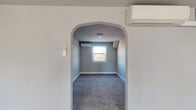 This single-floor space is thoughtfully laid out across several distinct areas, each finished in a cohesive, neutral gray color palette that creates a calm and clean atmosphere throughout. The entry opens into a bright, carpeted living area bathed in natural light from double windows, providing a welcoming and open feel. Adjacent rooms include multiple carpeted bedrooms, each featuring individual windows that invite natural light and views of the surrounding greenery. A distinctive arched doorway adds an architectural accent, leading to a secondary room equipped with its own wall-mounted mini-split unit for personalized climate control. The compact kitchen is outfitted with white cabinetry, a white range, and wood-look vinyl plank flooring, complemented by a large window overlooking lush outdoor scenery. The bathroom features white subway tile walling, wood-look vinyl plank flooring, a warm-toned wood vanity, and a window for natural ventilation. Mini-split heating and cooling units are installed in multiple rooms, ensuring year-round comfort. White trim and baseboards provide clean contrast throughout, while consistent flooring choices — plush carpet in living and sleeping areas, durable vinyl plank in wet areas — unify the overall design. The space presents a move-in-ready, functional layout suitable for a variety of occupants.
