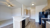 This single-floor residence showcases a clean, contemporary design with an open-concept layout that maximizes functionality and flow. The kitchen serves as a central feature of the space, outfitted with light wood cabinetry offering ample storage through upper and lower cabinets. Light-toned countertops provide generous workspace, complemented by a stainless steel sink and a black dishwasher tucked neatly beneath the counter. A stainless steel refrigerator anchors one end of the kitchen, while a peninsula-style breakfast bar extends outward, creating a natural transition into the adjoining living area. Warm wood-look flooring runs throughout, adding a sense of continuity and warmth to the space. The ceiling fan visible in the living area contributes to comfort and airflow across the open floor plan. Flush-mount ceiling lighting provides bright, even illumination throughout. Neutral white walls and a clean, minimalist aesthetic give the space a light and airy ambiance. A set of closet doors is visible toward the rear of the unit, indicating dedicated storage. Overall, the space offers a practical yet stylish environment well-suited for modern living.