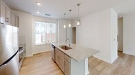 Located at 5421 Commonwealth Centre Pkwy in Midlothian, Virginia, this single-floor residence offers a clean, contemporary design with an open and airy layout. Light-toned hardwood-style flooring runs throughout the main living areas, complementing the crisp white walls and creating a cohesive, modern aesthetic.

The kitchen serves as a central highlight of the space, anchored by a large island with a granite countertop, an undermount sink, and stylish pendant lighting overhead. Gray shaker-style cabinetry lines the walls, paired with stainless steel appliances including a refrigerator, range, and microwave. Natural light pours in through a large window adjacent to the kitchen, brightening the open dining and living areas.

The living area is generously proportioned and features a ceiling fan, recessed lighting, and multiple windows with views of the surrounding landscape, allowing natural light to fill the room. A glass door provides access to an outdoor area, further enhancing the connection between interior and exterior spaces.

The bathrooms are well-appointed with granite-topped vanities, shaker-style cabinetry with brushed gold hardware, and contemporary light fixtures. One bathroom features a tub-shower combination with tile surround and a window for natural ventilation. Throughout the unit, the design prioritizes functionality and modern comfort with clean finishes and a neutral color palette.