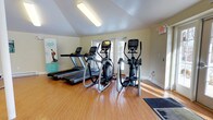 Located at 2991 W School House Ln in Philadelphia, PA, this fitness center offers a clean and functional workout environment designed to support an active lifestyle. The single-floor space is outfitted with multiple treadmills and elliptical cross-trainers, providing residents with a well-rounded selection of cardio equipment to meet their fitness needs. The room features warm hardwood-style flooring that adds a polished, welcoming feel to the space. Natural light filters in through a set of glass-panel doors along the back wall, creating a bright and energizing atmosphere ideal for exercise. The ceiling is finished with recessed fluorescent lighting, ensuring the space remains well-lit during all hours. Neutral-toned walls keep the environment feeling open and uncluttered, while motivational signage adds a touch of encouragement. An emergency exit sign is prominently displayed, reflecting attention to safety. The overall layout is thoughtfully organized to allow comfortable movement between equipment, making this fitness center a practical and inviting amenity.