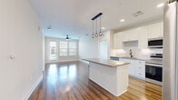 This single-floor, 2-bedroom, 2-bathroom residence in Ocoee, Florida offers a thoughtfully designed open-concept layout with contemporary finishes throughout. The kitchen serves as a centerpiece of the living space, featuring crisp white shaker cabinetry extending to the ceiling, a white subway tile backsplash, stainless steel appliances, a matte black undermount sink with a modern faucet, and a central island with a gray quartz-style countertop illuminated by stylish pendant globe lighting. Rich hardwood flooring flows seamlessly through the kitchen and living areas, which benefit from generous natural light and access to an outdoor balcony through large windows and a glass door, complemented by a ceiling fan overhead. Both bedrooms are carpeted in neutral tones and include ceiling fans, with the primary suite offering an en-suite bathroom and direct access to a walk-in closet area. The bathrooms are finished with white shaker vanities, gray countertops, matte black hardware, large mirrors, and warm hardwood-style flooring. The overall aesthetic is clean, modern, and bright, with a cohesive neutral palette that creates a welcoming, move-in-ready atmosphere throughout the entire residence.