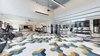 This well-appointed fitness center offers a generously sized, single-level layout divided into two functional zones. The main workout area commands attention with soaring double-height ceilings, dramatic suspended linear lighting, and ceiling fans that maintain airflow throughout the space. A bold geometric-patterned floor in blue, gold, and white anchors the room visually, creating a dynamic and energetic atmosphere. Horizontal blue stripes accent a feature wall, complementing the flooring and reinforcing the facility's cohesive design palette. Floor-to-ceiling mirrors line one wall, expanding the sense of space and providing visual feedback for users. Equipment is thoughtfully distributed and includes treadmills, cable machines, strength-training benches, rowing machines, and a dedicated free-weight and resistance band station. A blue open-cubby storage unit near the entrance provides organized storage for personal items.

A separate, glass-walled studio room offers a quieter training environment, finished with warm hardwood-style flooring and large windows that bring in natural light and outdoor views. This space is equipped with cycling bikes and rowing machines, making it well-suited for group or individual cardio sessions.
