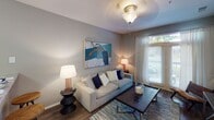 Located at 12201 S Tryon St in Charlotte, NC, this single-story unit offers a well-organized layout across several distinct living spaces. The living room features warm hardwood-style flooring, soft neutral gray walls, and a large window dressed with sheer white curtains that allow generous natural light to fill the room. A flush-mount ceiling fixture provides ambient lighting, and the space comfortably accommodates a full seating arrangement.

The kitchen is outfitted with light wood cabinetry, granite-style countertops, and white appliances including a range, microwave, and refrigerator. Dark hardwood-style flooring grounds the space, and a peninsula countertop adds functional workspace and a natural transition to adjacent areas.

Two bedrooms are carpeted and finished with the same cool gray wall tones found throughout the unit. Each room features a ceiling light fixture and windows that bring in natural light, creating a calm and restful atmosphere. The rooms offer comfortable space for standard bedroom furnishings.

The bathroom includes a light wood vanity with a granite-style countertop, a well-lit mirror with a multi-bulb fixture above, and hardwood-style flooring. A closet with sliding doors is conveniently positioned nearby. Overall, the unit presents a cohesive, neutral palette with consistent finishes across all rooms.