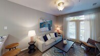 Located at 12201 S Tryon St in Charlotte, NC, this single-story unit offers a well-organized layout across several distinct living spaces. The living room features warm hardwood-style flooring, soft neutral gray walls, and a large window dressed with sheer white curtains that allow generous natural light to fill the room. A flush-mount ceiling fixture provides ambient lighting, and the space comfortably accommodates a full seating arrangement.

The kitchen is outfitted with light wood cabinetry, granite-style countertops, and white appliances including a range, microwave, and refrigerator. Dark hardwood-style flooring grounds the space, and a peninsula countertop adds functional workspace and a natural transition to adjacent areas.

Two bedrooms are carpeted and finished with the same cool gray wall tones found throughout the unit. Each room features a ceiling light fixture and windows that bring in natural light, creating a calm and restful atmosphere. The rooms offer comfortable space for standard bedroom furnishings.

The bathroom includes a light wood vanity with a granite-style countertop, a well-lit mirror with a multi-bulb fixture above, and hardwood-style flooring. A closet with sliding doors is conveniently positioned nearby. Overall, the unit presents a cohesive, neutral palette with consistent finishes across all rooms.