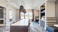 This single-floor co-working suite is thoughtfully designed to support both collaborative and focused work. The main conference area serves as the centerpiece, featuring a large white rectangular table surrounded by ergonomic leather chairs and illuminated by a trio of striking industrial-style pendant lights. Patterned carpet in earthy tones adds visual texture underfoot, while recessed lighting and natural light from large windows facing an outdoor courtyard view keep the space bright and welcoming. A built-in shelving and storage unit with navy cabinetry lines one wall, offering organized space for office supplies and equipment, while a cushioned bench nook provides an informal seating alternative. A private meeting room, accessible via a glass-paneled door, accommodates a smaller group around a second white conference table. This room features a wall-mounted flat-screen display, a warm wood-toned credenza, organic wall art accents, and an elegant multi-arm chandelier that adds a refined touch. The overall aesthetic is modern and professional, blending contemporary furnishings with warm materials to create an environment that is both functional and visually engaging — well-suited for meetings, focused work sessions, or team collaboration.