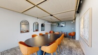 Located at 161 W 600 S in Salt Lake City, Utah, this single-floor conference room offers a well-appointed setting designed for professional meetings and collaborative sessions. The room centers around a large dark oval conference table surrounded by tan and amber upholstered chairs, providing comfortable seating for a sizable group. The ceiling features a distinctive grid of acoustic panels framed in dark trim, contributing both visual interest and sound management to the space.

A circular pendant light hangs above the table, offering focused illumination, while adjustable track lighting along the ceiling provides additional ambiance. One wall is adorned with three arched mirrors in black frames, adding depth and a contemporary design element alongside soft blue-gray paint. A large-scale abstract artwork hangs on an adjacent wall, complementing the room's refined aesthetic.

A wall-mounted flat-screen display and a wood-slat credenza with two lounge chairs define a secondary area suited for presentations or informal conversation. Floor-to-ceiling windows along one side of the room invite natural light and frame views of the Salt Lake City skyline and surrounding mountains. Patterned carpeting throughout ties the design elements together, creating a cohesive, modern atmosphere.