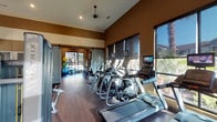 This fitness center, located along Meadow Ranch Pkwy in Richmond, TX, is a single-story facility designed to support a comprehensive workout experience. The space features soaring ceilings with pendant ceiling fans and warm earth-toned walls accented by a prominent exposed brick feature wall, creating an inviting yet functional atmosphere. Rich wood-look flooring runs throughout, adding warmth to the interior.

Large floor-to-ceiling windows line multiple walls, flooding the space with abundant natural light while offering views of an outdoor pool area and surrounding palm trees and landscaping. Adjustable roller shades provide light control when needed. Sliding barn-style doors offer a design-forward entry point to portions of the facility.

The cardio zone is well-stocked with a row of Matrix treadmills, ellipticals, stair climbers, upright and recumbent bikes, and spin cycles, each equipped with individual TV monitors. A dedicated strength training area includes cable machines, a multi-station rack system, a dumbbell rack, and weight benches, complemented by large wall mirrors and a flat-screen TV mounted on the brick wall. A corner nook features a built-in shelving unit stocked with complimentary bottled water and rolled towels, along with stability and exercise balls, rounding out the amenities of this well-appointed fitness center.